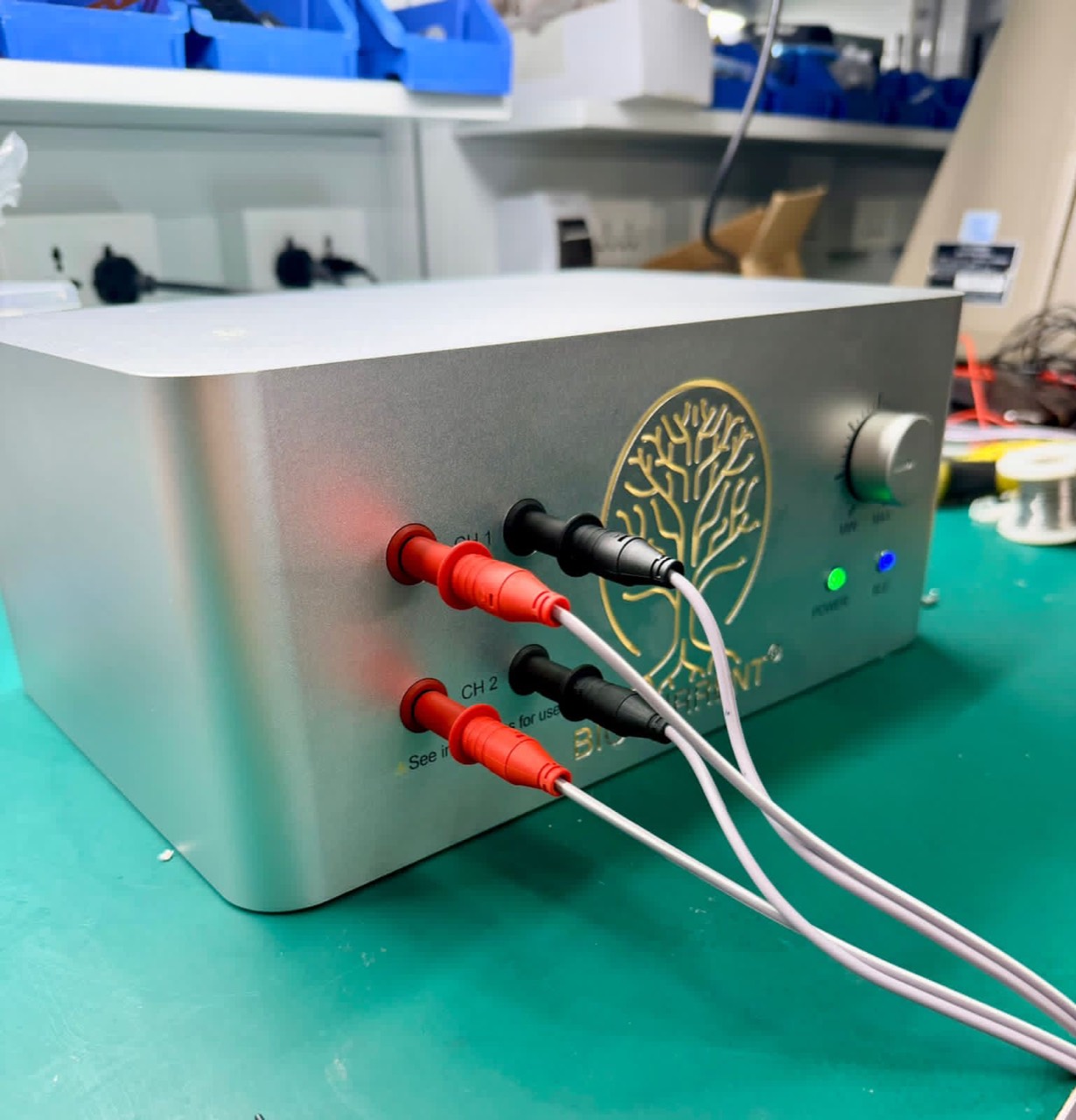 Actual Biocurrent V6 on a green workbench with two pairs of connected leads, a front-panel control, and illuminated indicators.